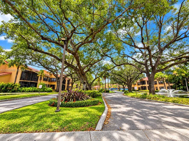 Landscaped grounds featuring mature canopy trees, manicured lawns, paved walkways, a water fountain, and stucco exterior buildings with tile roofs