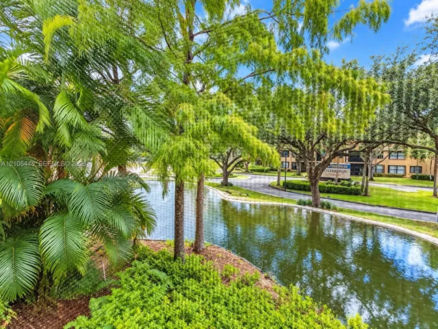 Waterfront grounds featuring mature canopy trees, manicured shrubs, and a paved walkway
