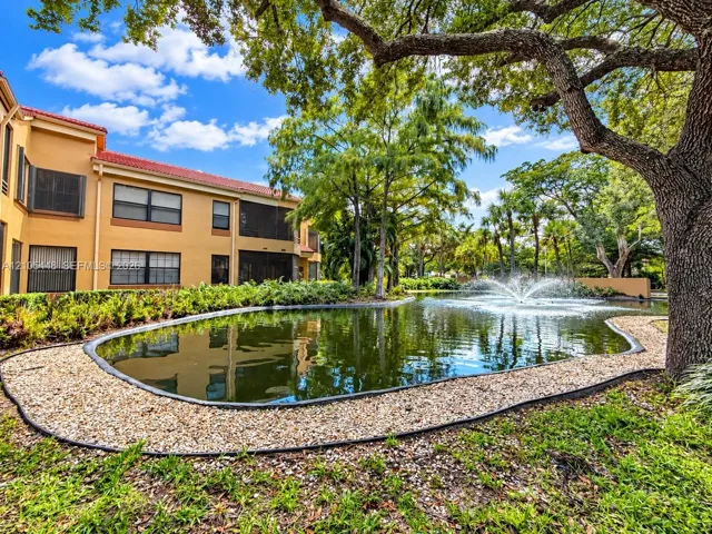 Multi-story exterior featuring tan stucco cladding, a red-tiled roof, screened-in lanais, a central water feature with a fountain, and mature landscaping