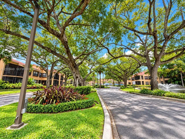 Paved driveway lined with mature canopy trees, manicured landscaping, water feature, and multi-story structures with screened lanais