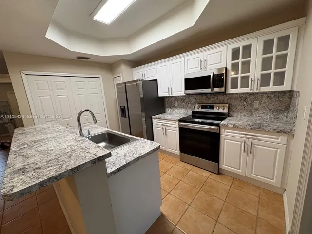 Kitchen featuring white cabinetry with some glass-front doors, stainless steel appliances, speckled countertops with a matching backsplash, and tile flooring