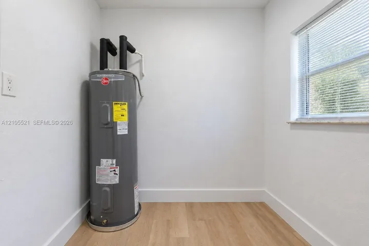 Water heater, wood-finish flooring, white walls, window with blinds, and white trim