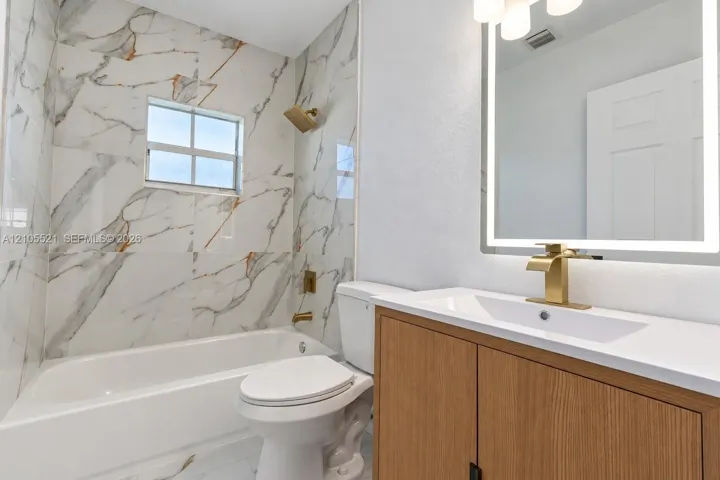 Bathroom featuring large format marble-look wall tile, a built-in bathtub, a vanity with wood-finish cabinetry, a white countertop, and a lighted mirror