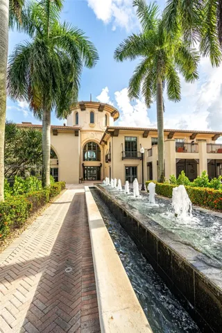 Stately stucco exterior with a prominent rotunda, a linear water feature with multiple fountains, a herringbone-patterned brick paver walkway, and mature palm trees