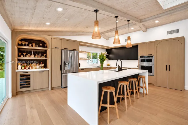 Vaulted wood-finish ceiling with exposed beams, wood-finish flooring, stainless steel appliances, built-in shelving with integrated lighting, and a kitchen island with a white countertop