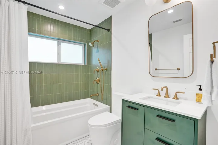 Bathtub with green vertical tile surround, gold-finish shower and bath fixtures, white vanity with green cabinetry, white countertop, and gold-finish faucet