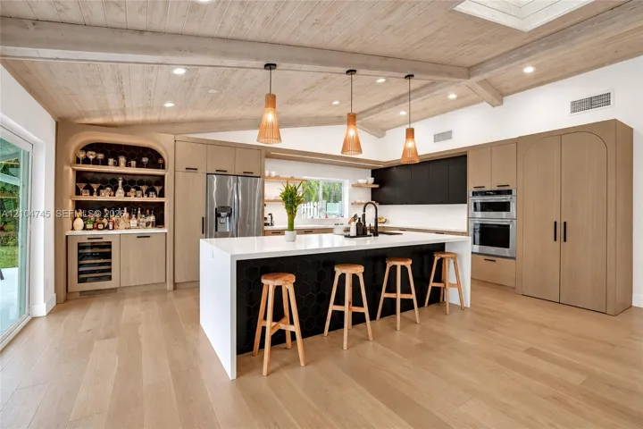 Wood-finish flooring, vaulted wood-paneled ceiling with exposed beams, built-in cabinetry with integrated appliances, kitchen island with hexagonal tile accent, and a built-in beverage center with a wine refrigerator