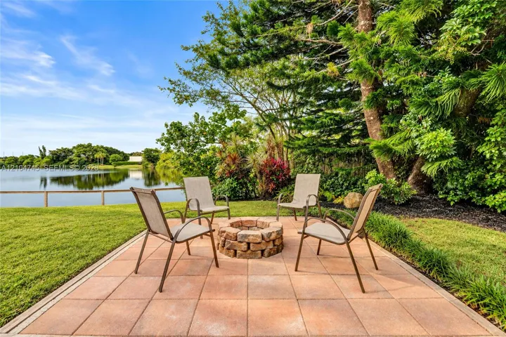 Paver patio with a stone fire pit, bordered by green lawn and diverse foliage, with a water feature in the background
