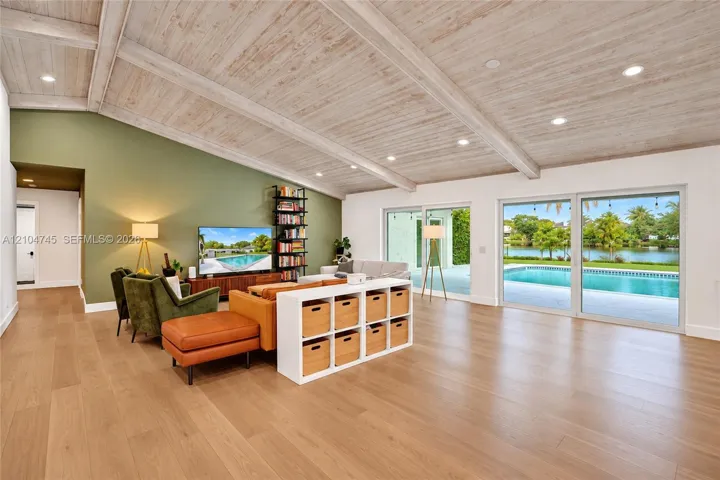 Vaulted ceiling with exposed wood beams, recessed lighting, wood-finish flooring, and expansive sliding glass doors opening to a private pool and waterway