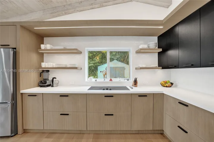 Integrated stainless steel refrigerator, light wood-finish lower cabinetry, white solid surface countertops, black upper cabinetry, and a built-in cooktop