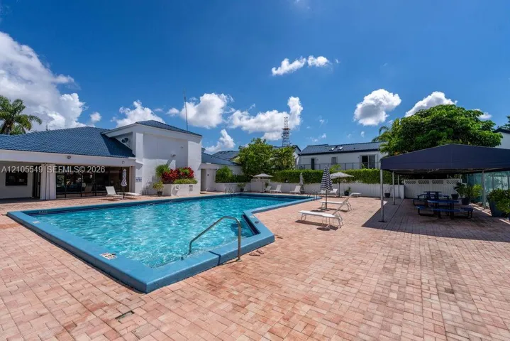 Expansive outdoor pool with a blue tile edge, surrounded by a large brick paver deck, permanent poolside canopy, and lush perimeter landscaping