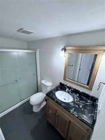 Frosted glass shower enclosure, dark large-format tile flooring, wood-finish vanity with an oval basin, integrated countertop, and framed wall mirror