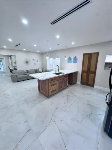 Spacious open-concept room featuring large format tile flooring, a kitchen island with a white countertop and integrated sink, pendant lighting, recessed ceiling lights, and wooden double doors