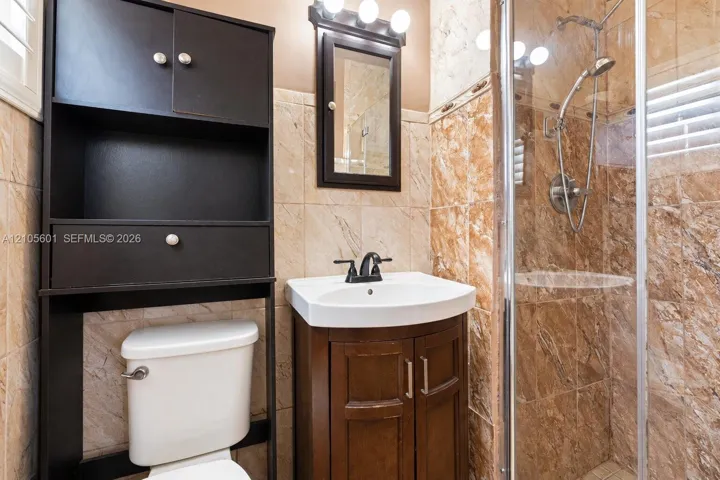 Glass-enclosed shower with a handheld shower fixture, wood-finish vanity with a white basin, dark wood-finish medicine cabinet, and tiled walls