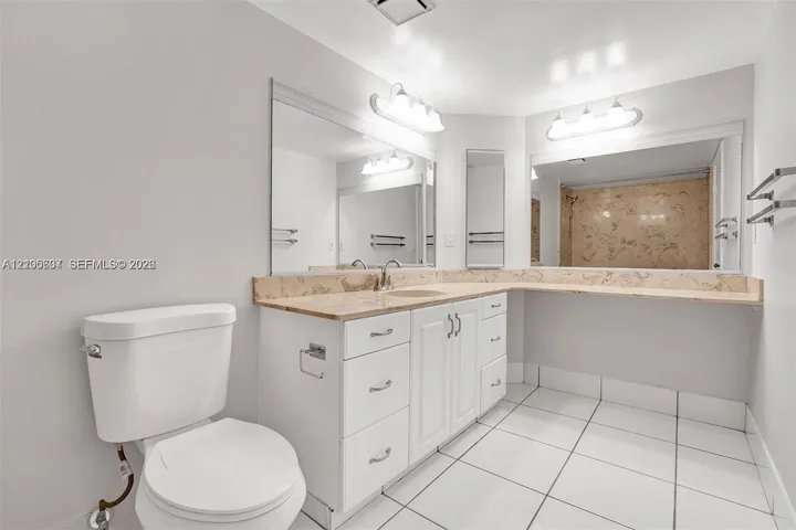 White tile flooring, white vanity with integrated sink, stone countertop, mirrored wall, and wall-mounted light fixtures