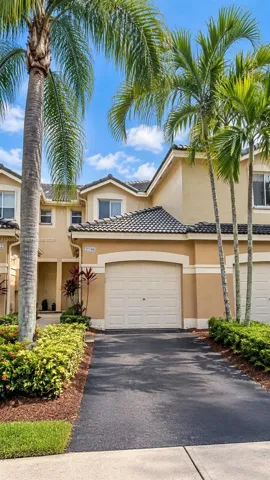 Stucco exterior with a tiled roof, arched garage entry, paved driveway, and established landscape plantings