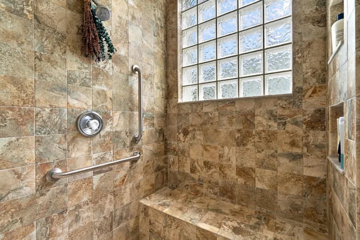 Tiled, primary walk-in shower with built-in bench, shower niche, multiple shower heads, and grab bars.