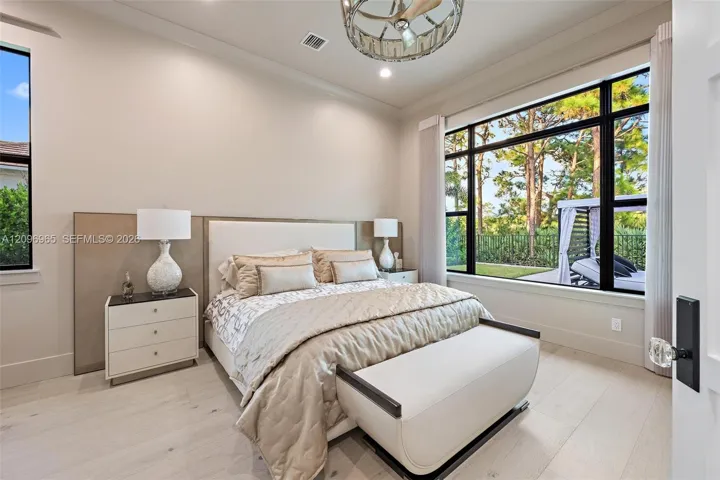 Light-toned wood-finish flooring, custom wall-panel headboard, expansive windows with black frames, and a tiered metal chandelier