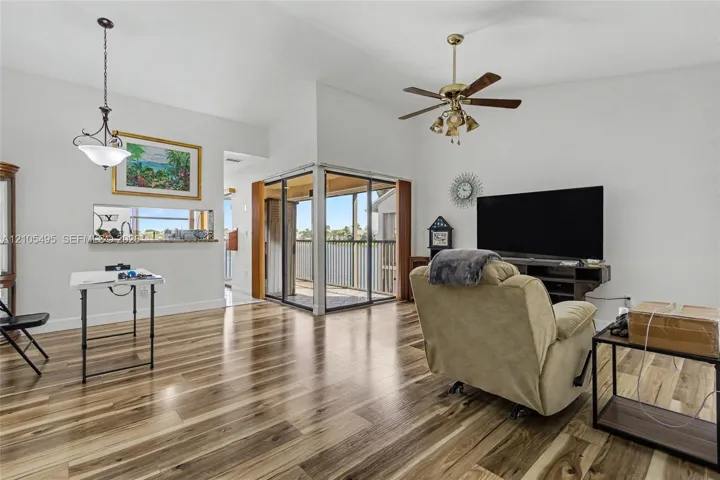 Living area featuring wood-finish flooring, vaulted ceilings, a ceiling fan with integrated light fixture, a pass-through opening, and sliding glass doors with access to an exterior screened balcony