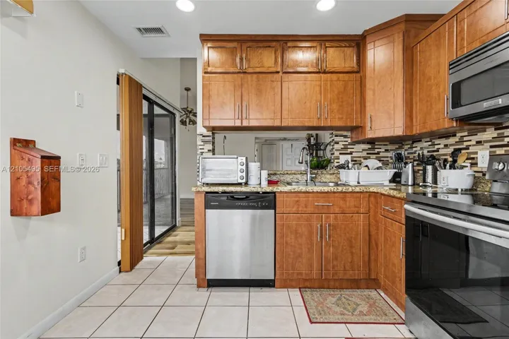 Kitchen featuring stained wood cabinetry, multi-tone tile backsplash, stone-finish countertops, stainless steel dishwasher, and ceramic tile flooring
