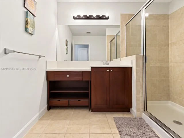 Bathroom vanity featuring a solid surface countertop, wood cabinetry with open lower shelving and drawers, full-width wall mirror, and a glass-enclosed shower with tile surround