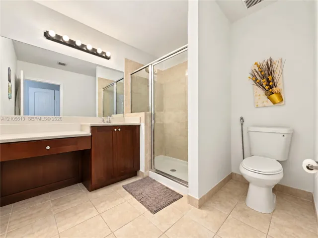 Bathroom featuring a wood-finish vanity with a light-toned countertop, a large wall-mounted mirror with multi-bulb light fixture, a glass-enclosed shower with tile surround, and tile flooring
