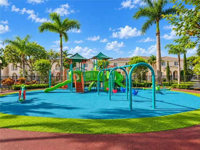 Playground facility featuring multiple slides, swing sets, and climbing structures on a multi-colored rubberized surface, surrounded by manicured landscaping and palm​​‌​​​​‌​​‌‌​‌‌​​​‌‌​‌​‌​‌​​​‌​​ trees