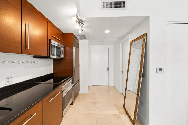 Galley kitchen with wood cabinetry, dark countertops, integrated stainless steel appliances, and tile flooring