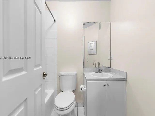 Bathroom featuring a vanity with a solid-surface countertop, an integrated sink, a wall-mounted mirror, a tub-shower combination with white tile surround, and a low-flow toilet