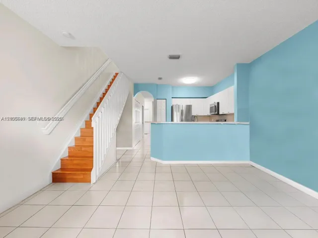 Open-concept interior featuring light-toned tile flooring, a staircase with wood-finish treads and white balusters, and a kitchen with white cabinetry and a breakfast bar