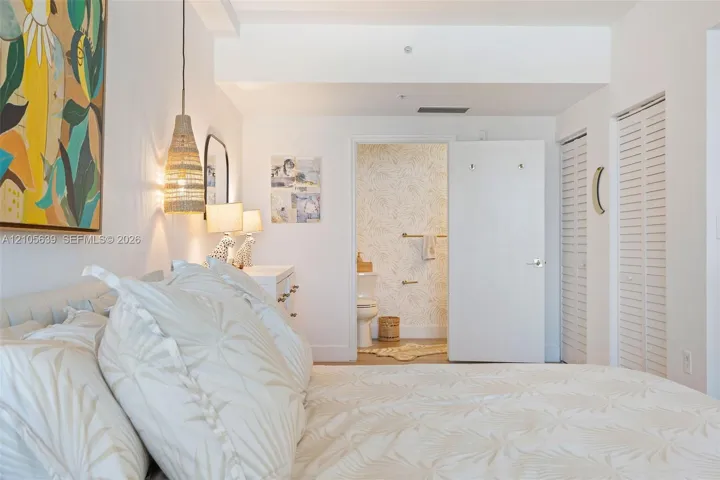 Neutral-toned bedroom featuring dual louvered-panel closets, a textured woven pendant light, and a dedicated washroom with patterned wallpaper