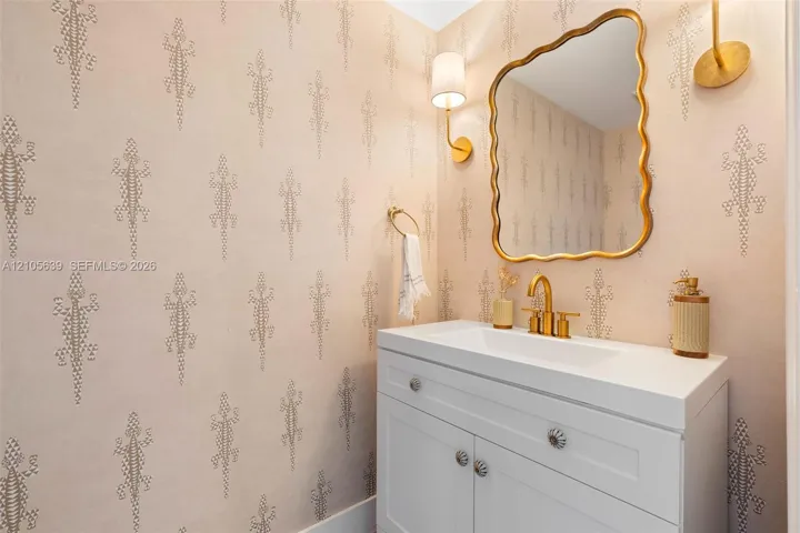 Patterned wall covering, white vanity cabinet with decorative hardware, white rectangular sink, gold-toned faucet and hardware, and wavy-edged mirror with gold-toned frame