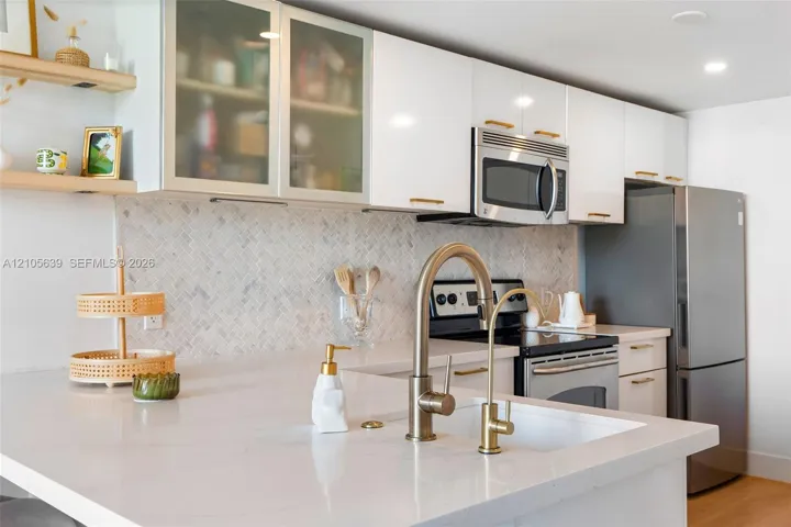 Kitchen featuring white cabinetry with glass-front uppers, herringbone-patterned stone backsplash, quartz countertops, brass-finish fixtures, and built-in stainless steel appliances