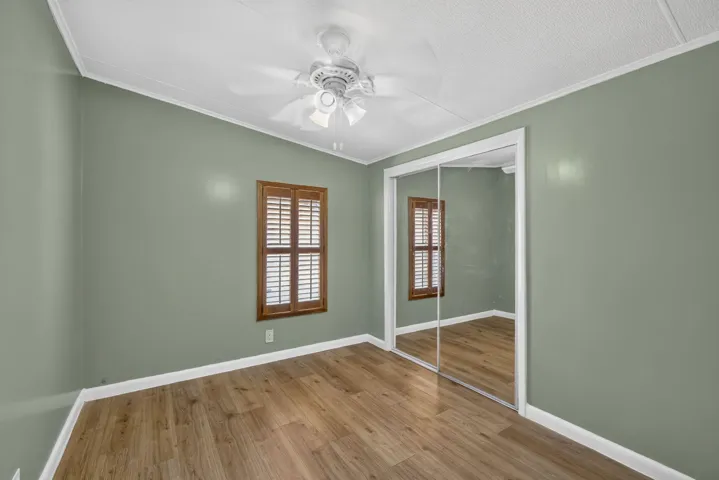 Wood-finish flooring, mirrored closet doors, window with wood shutters, ceiling fan, and white crown molding