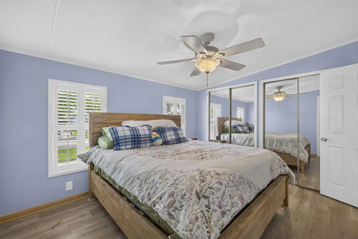 Wood-finish flooring, white window shutters, ceiling fan with light fixture, mirrored closet doors, and a paneled interior door