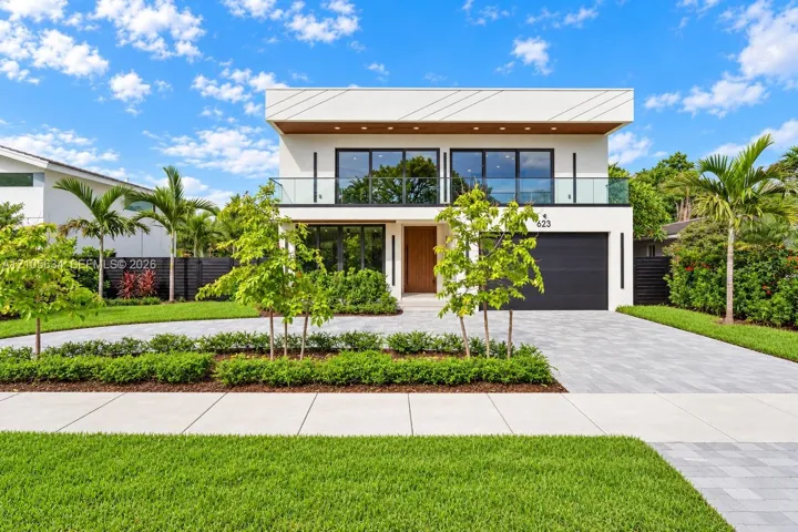 Modern two-story exterior featuring a flat roofline, upper-level glass-railing balcony, paver driveway, and wood-finish entry door