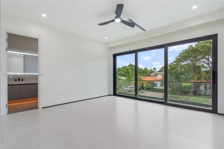 Large-format porcelain tile flooring, expansive floor-to-ceiling sliding glass doors with black frames, recessed LED lighting, three-blade ceiling fan, and balcony with glass railing