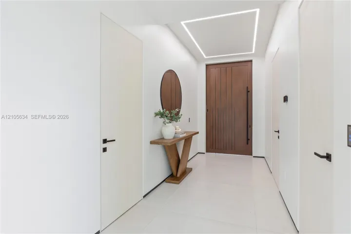 Entryway featuring a large-format wood-finish front door with a vertical pull handle, recessed linear ceiling lighting, minimalist flat-panel interior doors with matte black hardware, and large-format porcelain tile flooring