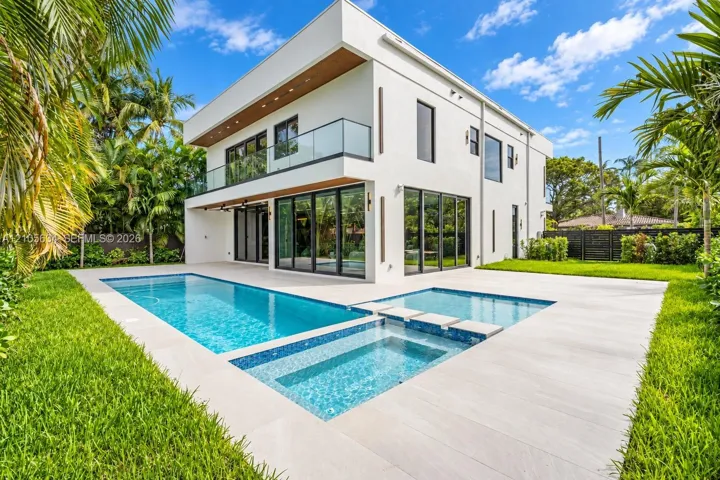 Modern two-story architectural exterior featuring smooth stucco finishes, floor-to-ceiling glass panel doors, a second-level glass-railed balcony, and an integrated swimming pool and spa surrounded by light-toned hardscaping