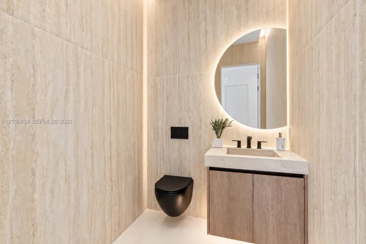 Full-height stone-finish wall tiling, wall-mounted matte black toilet, floating wood-finish vanity with integrated stone-finish basin, matte black widespread faucet, and semi-circular backlit wall mirror