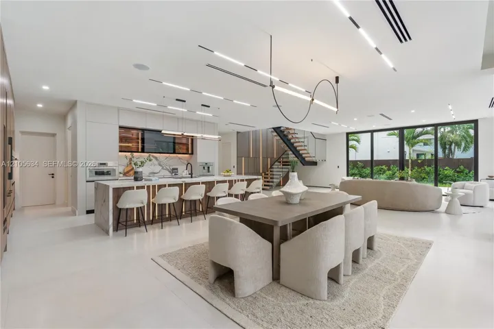 Open-concept interior featuring large-format flooring, a kitchen island with stone-surface counters, integrated LED accent lighting, floating staircase with metal railings, and floor-to-ceiling glass panel walls