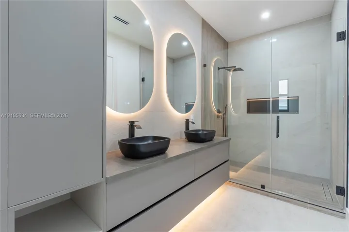 Floating dual-sink vanity with matte black fixtures, quartz-finish countertop, backlit oval mirrors, and a walk-in glass-enclosed shower featuring a built-in recessed​​‌​​​​‌​​‌‌​‌‌​​​‌‌​‌​‌​‌​​​‌​​ niche