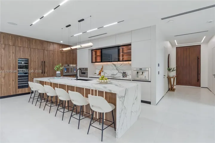 Modern kitchen featuring flat-panel wood-finish cabinetry, a large waterfall island with stone countertop, built-in stainless steel appliances, integrated under-cabinet lighting, and recessed linear ceiling illumination