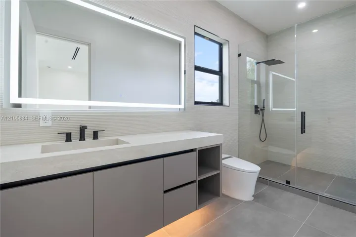 Floating vanity with integrated sink, stone-finish countertop, under-cabinet lighting, LED-backlit mirror, and glass-enclosed walk-in shower with matte black rainfall fixture