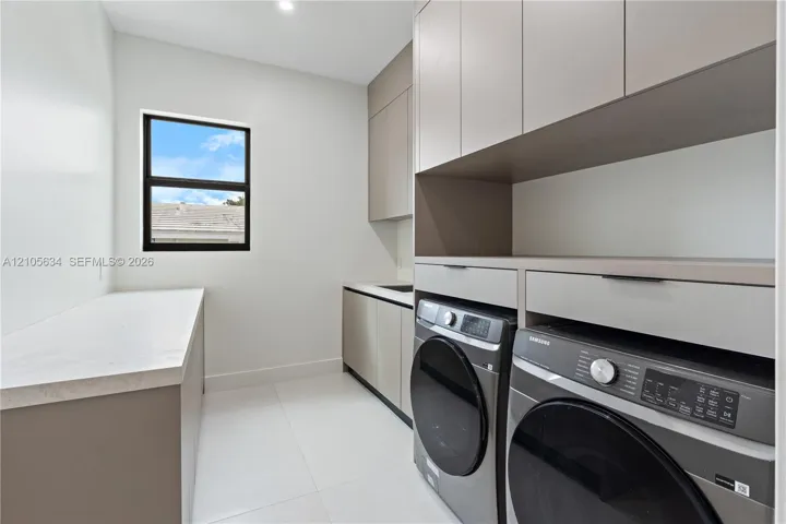 Laundry area featuring custom flat-panel cabinetry, light-toned stone-finish countertops, tile flooring, and integrated laundry appliances