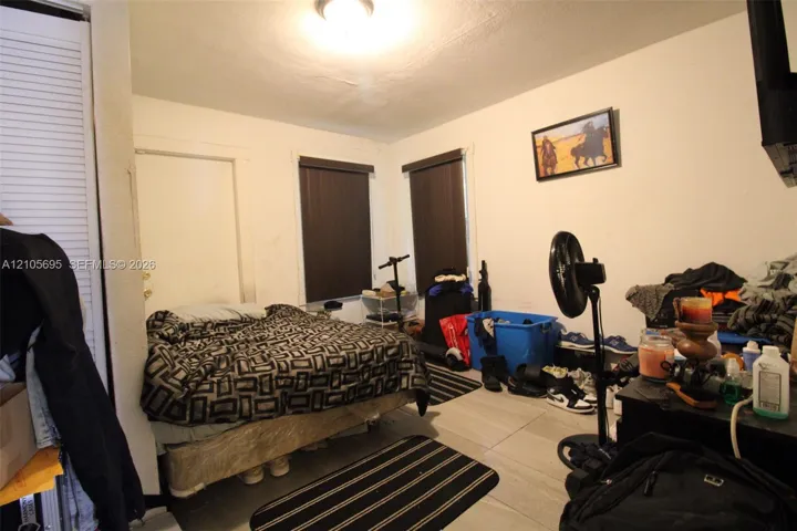 Interior room featuring large-format ceramic tile flooring, neutral wall finishes, dual windows with dark-tinted roller shades, and a closet with white louvered bi-fold doors
