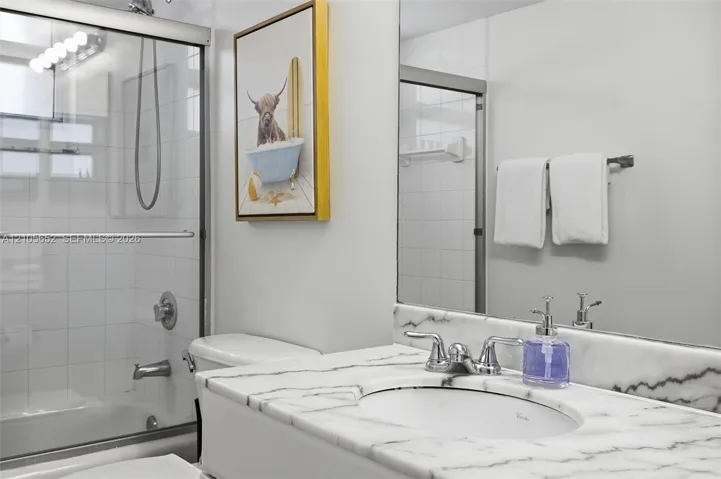 Bathroom featuring a vanity with a stone-surface countertop, an oval under-mount sink, a polished chrome faucet, a full-width mirror, and a tub-shower combination with a sliding glass enclosure and wall-mounted plumbing fixtures