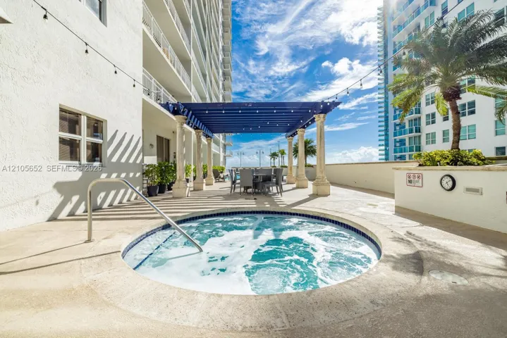 In-ground circular spa featuring a stainless steel handrail, shaded pergola with decorative stone columns, and overhead bistro-style lighting