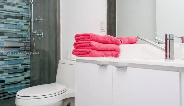 White vanity with a solid-surface countertop and chrome single-handle faucet, featuring a shower with a multi-colored glass tile accent wall and a chrome handheld shower head