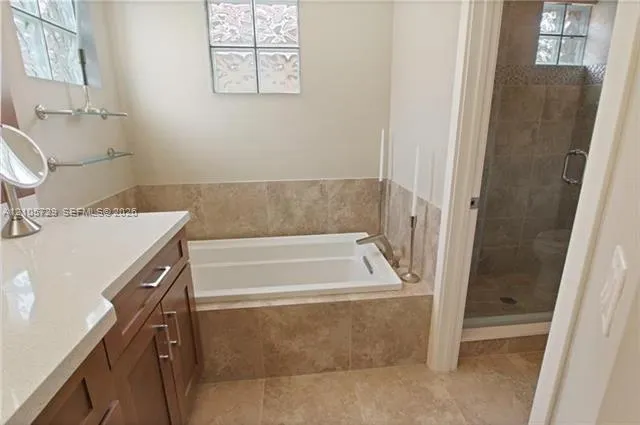Built-in soaking tub with stone-tile surround, vanity featuring a light-toned countertop, and separate glass-enclosed shower with tiled walls and flooring